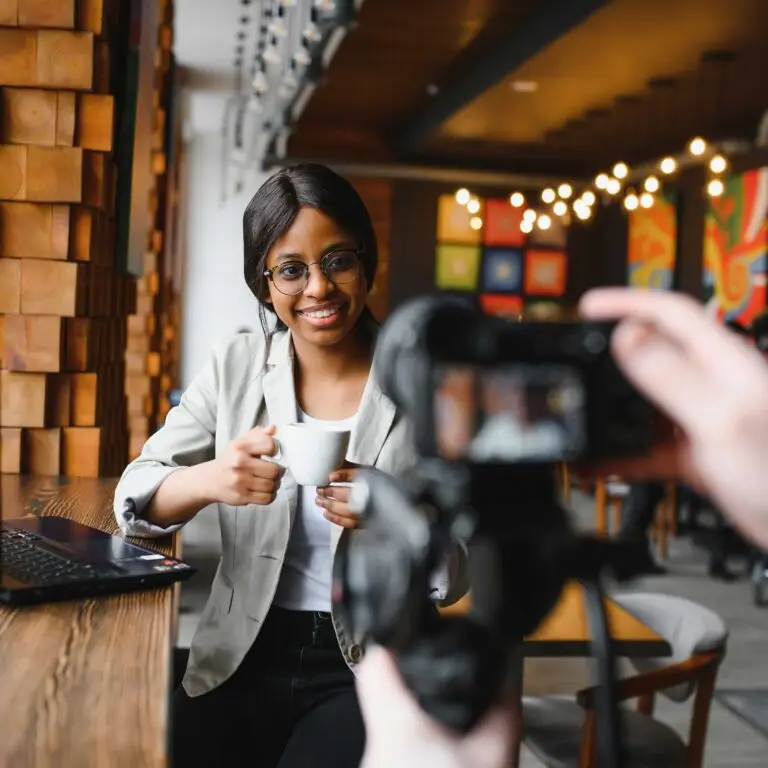 A woman being photographed at a café table with a coffee and laptop.