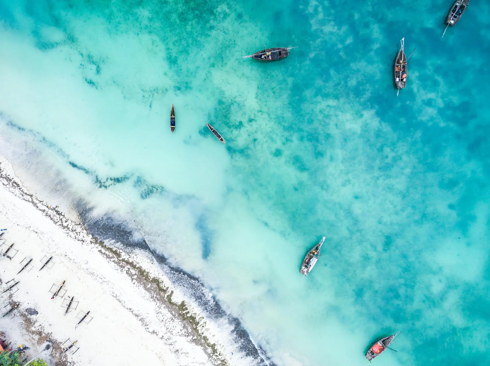 Aerial view of fishing boats in turquoise water beside a beach in Zanzibar.